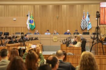 plenário com público em primeiro plano de costas e mesa alta composta pelas autoridades