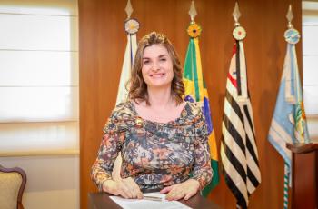 Juíza Andressa está no púlpito olhando para a foto, após assinar o termo de posse sobre o púlpito. Ao fundo, as bandeiras e o painel de madeira do Salão Nobre