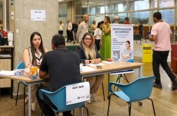 imagem de trabalhador negro sendo entrevistado por uma analista de recrutamento, ao lado de outra, que olha para a câmera. Todos sentados à mesa. Ao lado, um banner com os dizeres: Oportunidade de Emprego