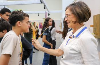 artista Michaela segura varetas de difusor para adolescente sentir o aroma. Ao fundo, outros adolescentes interagem com as obras