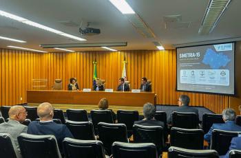 À frente da sala, quatro autoridades estão sentadas atrás de uma mesa de madeira, diante das bandeiras do Brasil e do Estado de São Paulo. À direita, um telão exibe slide do projeto apresentado pelo TRT-15. Na plateia, participantes acompanham a exposição sentados em cadeiras pretas dispostas em fileiras. O ambiente possui iluminação clara e paredes revestidas em madeira.