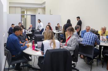 A foto retrata um ambiente com mesas e várias pessoas. Estão participando de audiências conciliatórias.