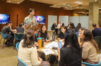 Instrutor está de pé falando com laboratoristas em atividades, agrupados em mesas redondas