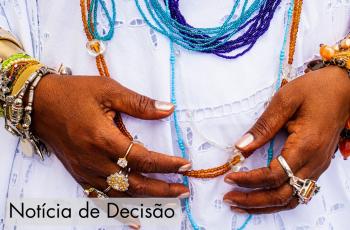 Imagem em close das mãos de uma pessoa de pele negra posicionadas à frente do corpo, segurando delicadamente um colar de contas em tons de âmbar. A pessoa veste uma roupa branca rendada e usa diversos colares de miçangas sobrepostos, em tons de azul, turquesa, roxo, branco e âmbar. Nos dois pulsos há várias pulseiras metálicas e de miçangas coloridas, e em ambas as mãos há diversos anéis prateados e dourados. O fundo é preenchido pela própria vestimenta branca, destacando as cores vibrantes dos acessórios. 