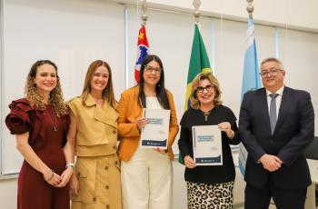 "Foto colorida, em ambiente institucional. Cinco pessoas estão lado a lado, sendo quatro mulheres e um homem, todos em pé e sorrindo para a câmera. Duas das mulheres, posicionadas ao centro, seguram exemplares de uma publicação. Ao fundo, há bandeiras em mastros, incluindo a do Brasil e outras bandeiras institucionais, além de grandes janelas com persianas claras. A imagem transmite um momento de registro oficial de entrega ou lançamento de material."