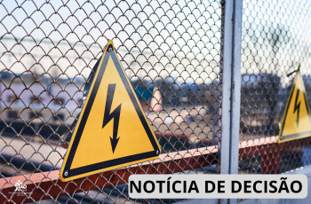 : Ilustração mostra, em primeiro plano, uma placa triangular amarela de advertência para risco de choque elétrico, com borda preta e símbolo de um raio, afixada em uma cerca de tela metálica. Ao fundo, desfocados, aparecem estruturas e equipamentos de uma área industrial ou subestação elétrica, além de uma segunda placa de advertência semelhante. No canto inferior direito, há a inscrição "NOTÍCIA DE DECISÃO" sobre uma faixa cinza. No canto inferior esquerdo, aparece a marca comemorativa dos 40 anos do TRT-1