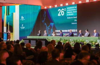 A imagem mostra um grande auditório durante um congresso jurídico, visto a partir da plateia.  No palco, há uma longa mesa revestida de verde, decorada na parte frontal com arranjos de plantas. Oito pessoas estão sentadas atrás da mesa, aparentemente compondo uma mesa de autoridades ou palestrantes. À esquerda do palco, uma mulher está em pé diante de um púlpito, aparentemente falando ao público.  Ao fundo, grandes painéis de LED exibem uma identidade visual em tons de azul, turquesa e verde, com elementos 