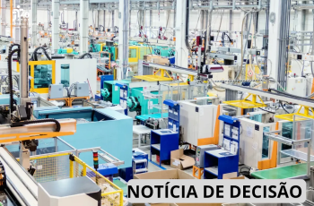 Fotografia de uma área industrial, com diversas máquinas e equipamentos de produção distribuídos pelo amplo espaço. Há estruturas metálicas, tubulações, cabos, esteiras, painéis e equipamentos de proteção em cores predominantemente azul, verde, amarelo, branco e laranja. Ao fundo, observam-se outras máquinas e linhas de produção. Na parte inferior direita, há uma faixa branca com a inscrição “NOTÍCIA DE DECISÃO”