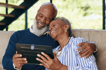 Foto de dois idosos abraçados sentados. Eles seguram um tablet e sorriem.